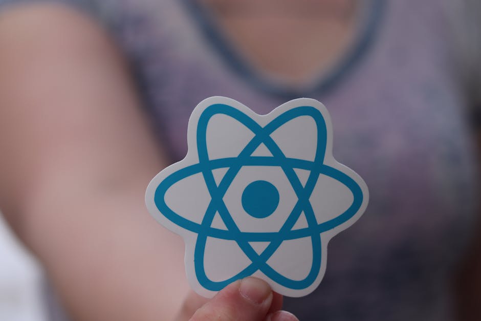 Close-up of a person holding a sticker featuring a blue atom symbol, with a blurred background showing a person in casual attire. The setting appears to be indoors, with natural lighting illuminating