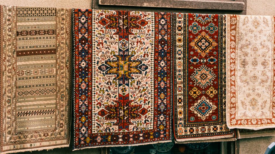 A collection of intricately patterned Persian and Oriental rugs draped over a wooden furniture piece in a well-lit room, with some rugs hanging down towards a tiled floor. The rugs feature vibrant col